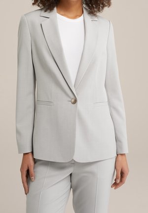 Kvinde iført en lysegrå skræddersyet blazer med en enkelt lukket knap, kombineret med matchende bukser og en hvid top under.
