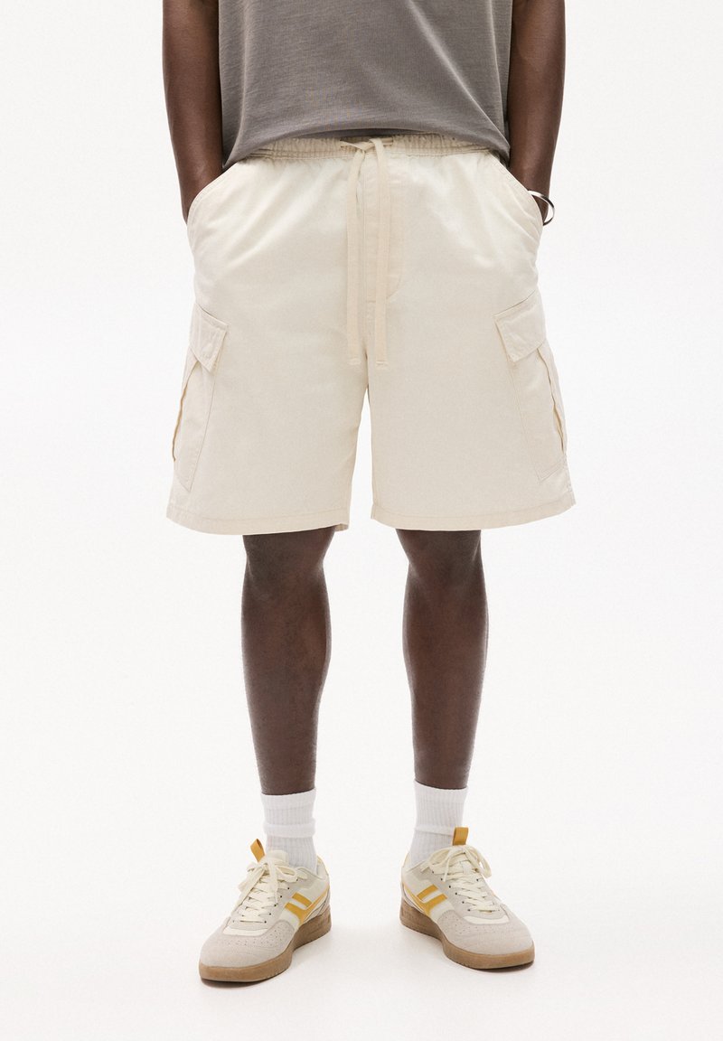 Kremowe szorty cargo z elastycznym paskiem, bocznymi kieszeniami i luźnym krojem. Noszone z jasnymi tenisówkami i białymi skarpetkami. Gładka szara koszulka.