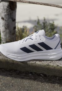 White athletic shoe with navy three stripes, textured upper, and thick sole placed on a stone ledge outdoors near wooden post and rope.