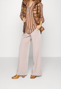 Veste matelassée marron avec fermeture éclair, blouse rayée marron et blanche, pantalon large crème, et sandales à talons marron. Matières lisses et texturées.