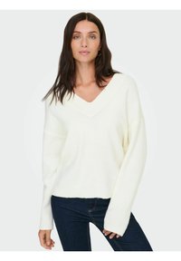 Pull blanc en tricot avec col en V, épaules tombantes et ourlet côtelé. Porté sur un jean foncé, il présente une texture douce et une coupe décontractée.