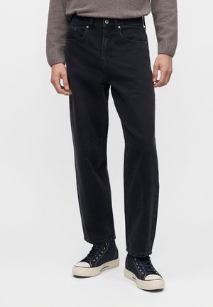 Personne portant un jean noir droit, des baskets montantes bleu marine avec semelles blanches et un pull gris, debout devant un fond uni.