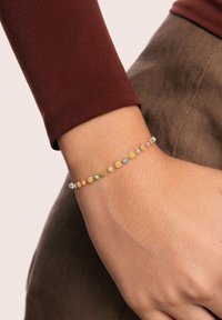 Handgelenk, das ein zartes Goldkettchen mit kleinen, mehrfarbigen Perlen trägt, kombiniert mit einem braunen Langarmshirt und taupefarbenen Hosen.