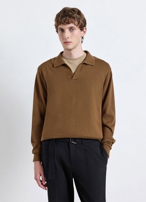 Junger Mann mit hellbraunen lockigen Haaren, der einen braunen Langarmpullover über einem beigen Hemd und schwarze Hose mit Gürtel trägt, steht vor weißem Hintergrund.