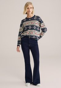 Svetr s vícebarevnými geometrickými vzory v krémové, námořnické, červené a zelené barvě; spárovaný s tmavě modrými zvonovými džíny; model má na sobě světlou obuv.