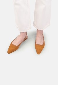 Picioare în pantaloni albi purtând pantofi slingback din suede bej cu vârful ascuțit pe un fundal alb.
