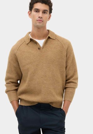 Junger Mann trägt einen hellbraunen gerippten Strickpullover mit Kragen und Knöpfen, weißes Unterhemd und dunkle Hose, Hände in den Taschen.
