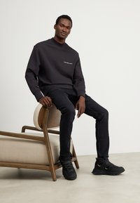 Felpa nera a girocollo con logo "Calvin Klein Jeans", abbinata a jeans scuri e stivali neri con lacci. Modello seduto su una sedia di legno.