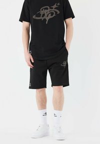 Black t-shirt and black shorts featuring a large gray logo. White socks with black accents and white sneakers complete the outfit.