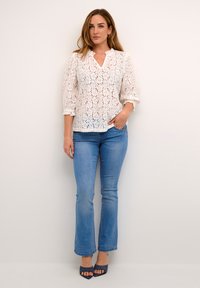 Blouse en dentelle blanche avec des manches trois-quarts, décolleté en V, motif floral, associée à un jean flare bleu clair et des escarpins ouverts marine.