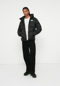 Jeune homme avec des cheveux tressés portant une veste rembourrée noire, un t-shirt blanc, un pantalon noir et des baskets noires se tenant devant un fond blanc.
