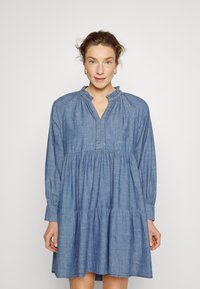 Vestido de ganga com gola em babados, mangas longas e saia em camadas. O tecido é suave, de cor azul clara e com um corte descontraído.