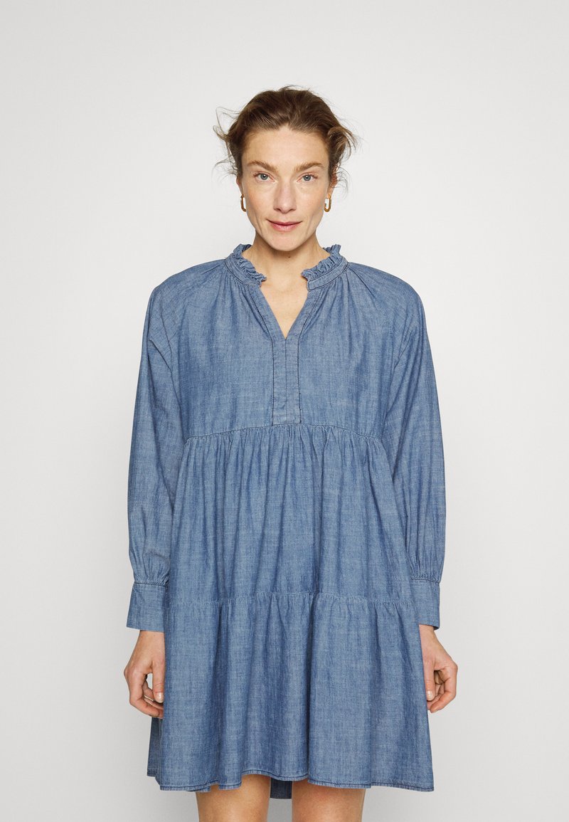 Vestido de ganga com gola em babados, mangas longas e saia em camadas. O tecido é suave, de cor azul clara e com um corte descontraído.