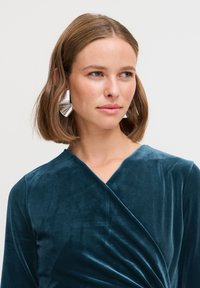 Teal sammetklänning med omslag design och långa ärmar, med V-ringning. Silverfärgade geometriska örhängen fulländar looken.
