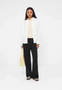 Bílé košilové tričko, béžový top, černé široké kalhoty, černé sandály s dekorativními akcenty, držící světle žlutou tašku. Minimalistický styl.