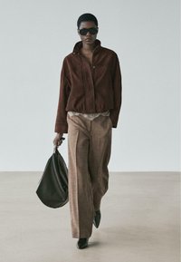 Eine braune Wildlederjacke mit hohem Kragen, kombiniert mit beige karierten, weit geschnittenen Hosen und schwarzen spitzen Schuhen, dazu eine schwarze Tasche in der Hand.