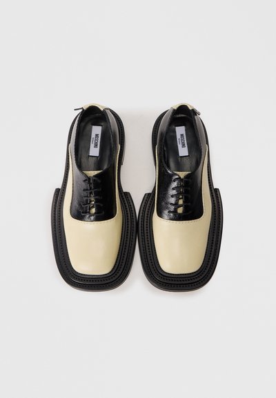 Chaussures en cuir noires et crème avec un bout arrondi, un système de laçage et une semelle épaisse noire. Présente des textures contrastantes et des détails de couture.