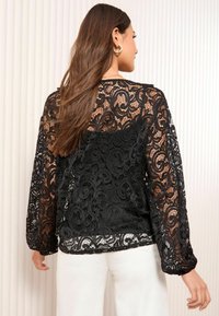 Blouse en dentelle noire présentant des motifs en spirale délicats, des manches longues et une texture transparente. L'ourlet est légèrement courbé et froncé aux poignets.