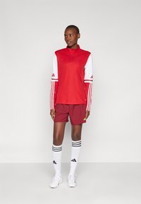 Rød atletisk zip-up top med hvide ærmer, parret med maroon shorts. Hvide sportsstrømper med sorte striber og hvide fodboldstøvler.