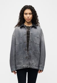 Γυναίκα με σγουρά μαλλιά φορώντας ένα φαρδύ ξεθωριασμένο τζιν μπουφάν πάνω από ένα μαύρο τοπ και μαύρο παντελόνι, που στέκεται μπροστά σε ένα απλό ανοιχτό φόντο.