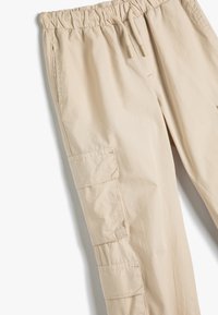 Beige Cargo-Shorts mit elastischem Bund und Kordelzug. Merkmale sind seitliche Taschen und eine aufgesetzte Tasche am linken Bein. Glatte Textur.