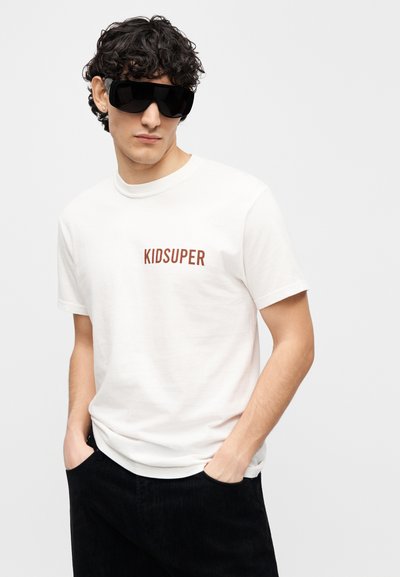 Λευκό βαμβακερό μπλουζάκι με λογότυπο "KIDSUPER" σε καφέ, σε συνδυασμό με σκούρο παντελόνι κοτλέ. Το μοντέλο φοράει μεγάλα μαύρα γυαλιά ηλίου.