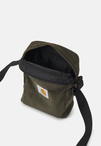 Pieni oliivinvihreä corduroy-olkalaukku, jossa on musta säädettävä hihna, avoin vetoketju ja Carhartt-logopatchi edessä.