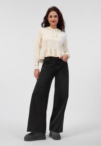 Felpa corta di colore crema chiaro con orlo arricciato, abbinata a jeans a gamba larga neri e scarpe con piattaforma nere. Tessuto liscio con una morbida consistenza.