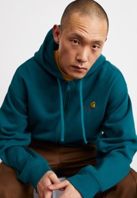 Mėlynos zip-up džinsinės striukės su raišteliais, su maža raudona logo ant krūtinės, derinamos su rudos spalvos kelnėmis. Minkšta medžiaga, laisvas prigludimas.