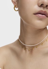 Pendientes de aro de oro, collares de perlas en capas con charms de oro. Acabado texturizado y brillante con colgantes de corazón y estrella, primer plano sobre la piel.