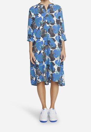 Femme porte une robe midi blanche avec de grands imprimés floraux bleus et noirs, des manches trois-quarts et des baskets blanches avec des semelles bleues.