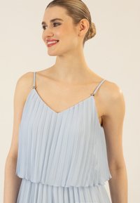 Vestido plisado de color azul claro con escote en V y tirantes finos adornados con pequeños detalles de plata. Tela suave y fluida.