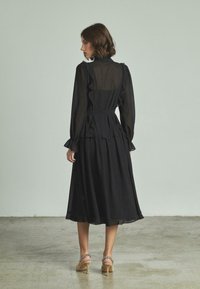 Un vestido negro con cuello alto, tejido transparente, acentos de volantes y una falda acampanada, combinado con sandalias de tiras beige.