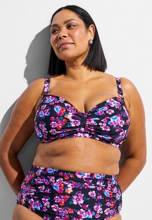 Žena so stredným odtieňom pokožky, ktorá má na sebe čierne kvetinové bikiny s vysokým pásom, stojí pri bielej stene s rukami za chrbtom.