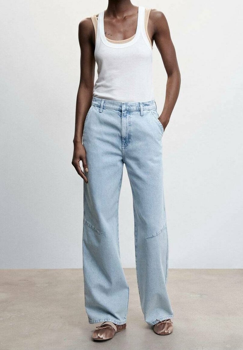 Personne portant un débardeur blanc, un jean bleu clair à jambes larges avec des poches avant et des ballerines en satin beige, debout avec une main dans la poche.