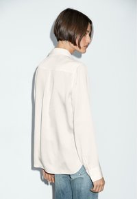 Long-sleeve, cream blouse with a buttoned collar and a back yoke detail, paired with light blue denim jeans. Smooth texture, minimal design.