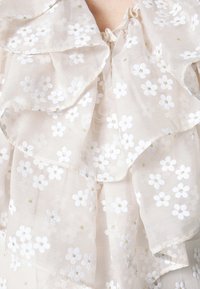Gros plan sur un tissu beige transparent avec broderie florale blanche et centres en fil d'or, superposés en volants sur la peau.