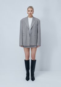 Blazer grigio oversize con rever a punta e due bottoni, abbinato a una gonna corta grigia e stivali neri in pelle con punta, alti fino al ginocchio.