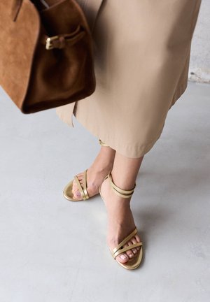 Personne portant une jupe beige et des sandales dorées à brides, tenant un sac à main en daim marron, debout sur un sol gris clair.