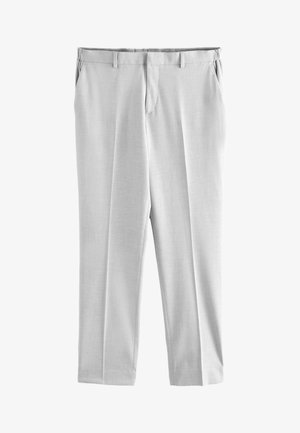 Mężczyzna stojący, ubrany w jasnoszary garnitur, białą koszulę, poszetkę oraz brązowe eleganckie buty na jednolitym jasnym tle.