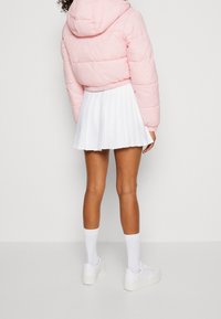 Person som bär en rosa huva-pufferjacka, vit plisserad kjol, vita strumpor och vita sneakers, som står mot en enkel vit bakgrund.