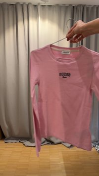 Käsi hoiab metallist riidepuud, millel ripub pikkade varrukatega roosa särk, mille rinnal on tikitud kiri "GUESS jeans", taustal beežid kardinad.