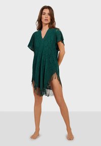 Cubierta de encaje verde con escote en V, mangas cortas y dobladillo con flecos. Presenta un patrón floral y es semitransparente.