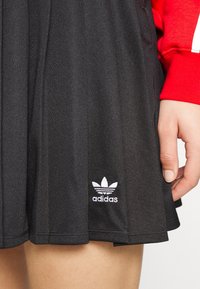 Czarna spódnica sportowa wykonana z teksturowanej tkaniny, z plisami i haftowanym białym logo Adidas w pobliżu hemu.