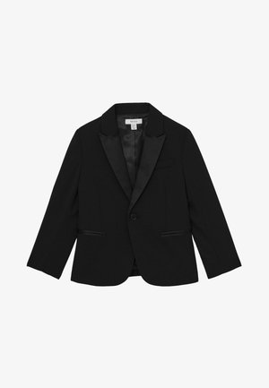 Barn med krøllet brunt hår iført en sort blazer og matchende bukser, står med ryggen mod et ensfarvet lyst baggrund.