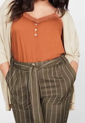 Frau trägt ein verbrannt oranges Camisole mit Spitzenbesatz, eine beige Strickjacke und eine hochtaillierte olivgrüne Hose mit weißen vertikalen Streifen und Stoffgürtel.