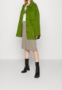 Giacca di lana verde con colletto e grandi tasche frontali, abbinata a una gonna midi a quadri beige e stivali neri con tacco a blocco.