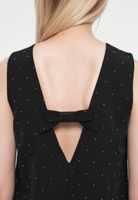 Rochie neagră fără mâneci, cu un decupaj în formă de V la spate, împodobită cu o mică fundiță neagră și puncte argintii dispersate pentru ornament.