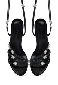 Sandales en cuir noir avec de fines lanières, ornées de petits clous en forme de rayons de soleil en argent, dotées de semelles de marque et de lacets autour de la cheville.