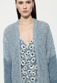 Hellblauer, gestrickter Cardigan mit grober Textur, offener Vorderseite und weiten Ärmeln, getragen über einem Blumenkleid in Blau- und Weißtönen.